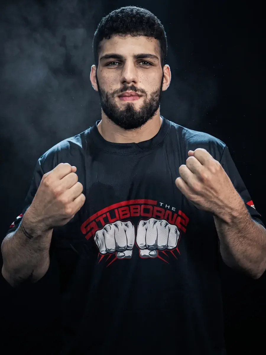 Coach Hadi Almir in a Stubborns shirt, fists up in guard against a dark, smoke-lit backdrop