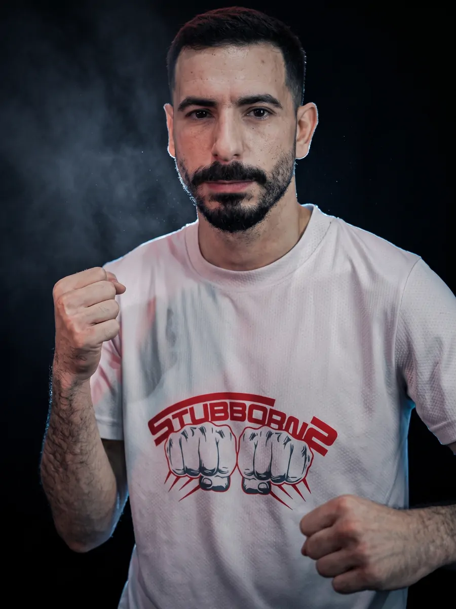 Head coach Hussein Ghaddar in a Stubborns shirt, fists up in guard against a dark, smoke-lit backdrop